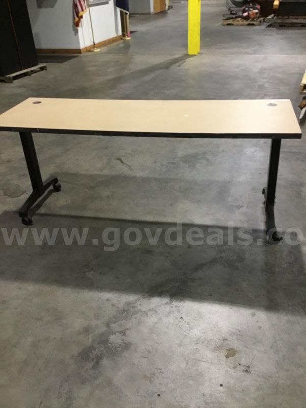 72 x 24 light tan rectangular table; with two black leg stands “DH”